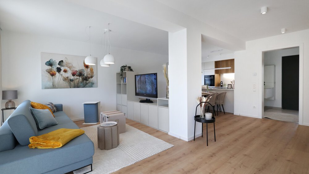 Modernes offenes Wohnzimmer mit blauem Sofa, Wandkunst, Fernseher und Küchenbereich mit Holzböden und weißen Wänden.