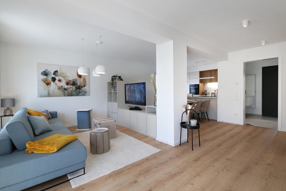 Modernes offenes Wohnzimmer mit blauem Sofa, Wandkunst, Fernseher und Küchenbereich mit Holzböden und weißen Wänden.