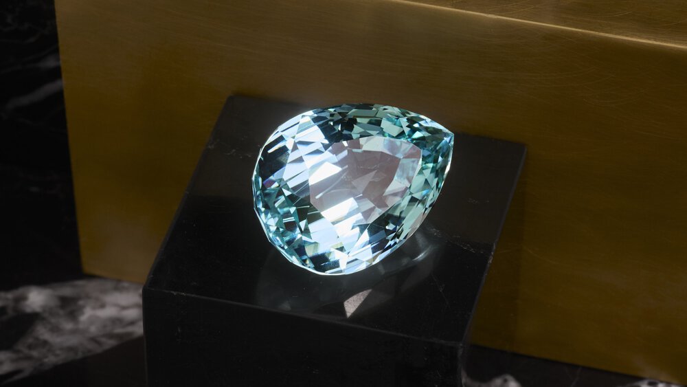A large, pear-shaped aquamarine gemstone on a black base against a golden background.