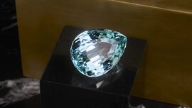 Ein großer, birnenförmiger Aquamarin-Edelstein auf einem schwarzen Sockel vor einem goldenen Hintergrund.