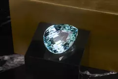 Ein großer, birnenförmiger Aquamarin-Edelstein auf einem schwarzen Sockel vor einem goldenen Hintergrund.