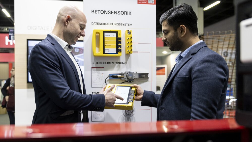 Zwei Männer in Anzügen untersuchen einen Betonsensor an einem Stand einer Technologieausstellung.