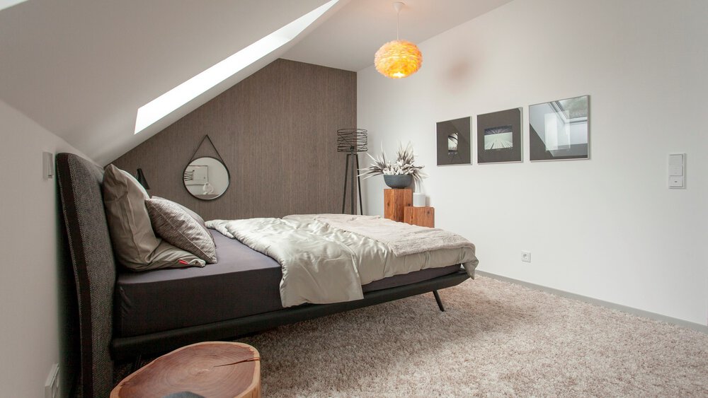 Modernes Dachgeschossschlafzimmer mit Dachschräge, Doppelbett, Kunstwerken an den Wänden und einem kleinen Nachttisch aus Holz mit Lampe.