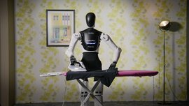 A humanoid robot irons a black shirt on an ironing board in a room with floral wallpaper and a framed picture.