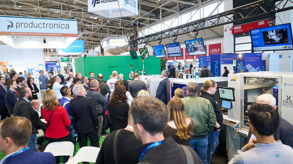 Eine gut besuchte Veranstaltungshalle der productronica Messe mit zahlreichen bunten Ständen, mit Werbetafeln, Monitoren und Beispielmaschinen.