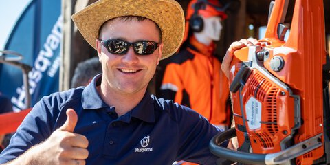 Man in a dark blue shirt, straw hat and sunglasses poses next to an orange chainsaw and raises his thumb in the air.
