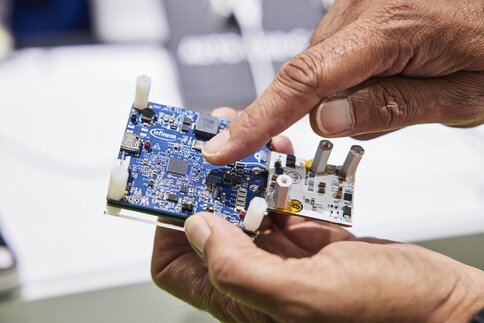 A person holds a blue circuit board with electronic components and points to it.