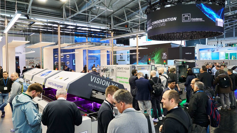Blick in eine gut besuchte Messehalle mit zahlreichen bunten Ständen, aufgeklappten Maschinen und Automaten und Werbebannern über den Köpfen der Besucher.