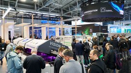 Blick in eine gut besuchte Messehalle mit zahlreichen bunten Ständen, aufgeklappten Maschinen und Automaten und Werbebannern über den Köpfen der Besucher.