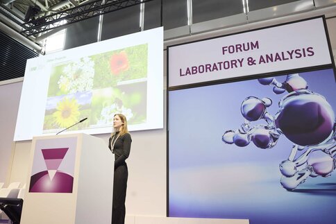 A woman stands behind a lectern. Behind her is a presentation with flowers and an analytica sign with the inscription 
