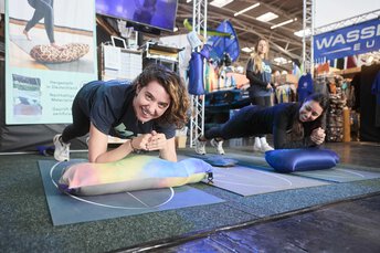 Zwei Frauen sind in Plank-Position mit den Unterarmen auf einem Kissen abgestützt. Beide lächeln in die Kamera.