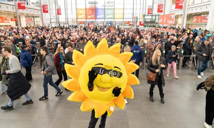 Das Maskottchen der f.re.e, eine gelbe Sonne mit Sonnenbrille, steht im Eingangsbereich der Messe und posiert vor den Besuchern an den Ticketkontrollen.