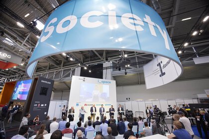 Panel discussion with 6 participants in a large exhibition hall, under a large 