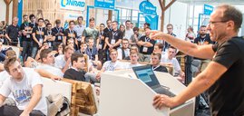 Ein Redner spricht zu einem sitzenden Publikum bei einer Konferenz oder einem Tech-Event in geschlossenen Räumen mit vielen Teilnehmern.