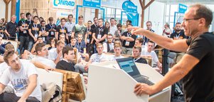 A speaker addresses a seated audience at a conference or a tech event in closed rooms with many participants.