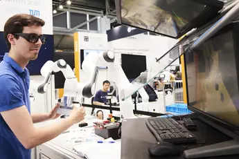 A person with dark glasses interacts with a robotic arm at a technology stand and looks at a screen.