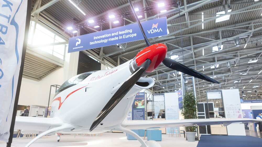 Ein weiß-rotes leichtes Propellerflugzeug steht in einer Halle der productronica unter dem Schild „Innovation and leading technology made in Europe“.