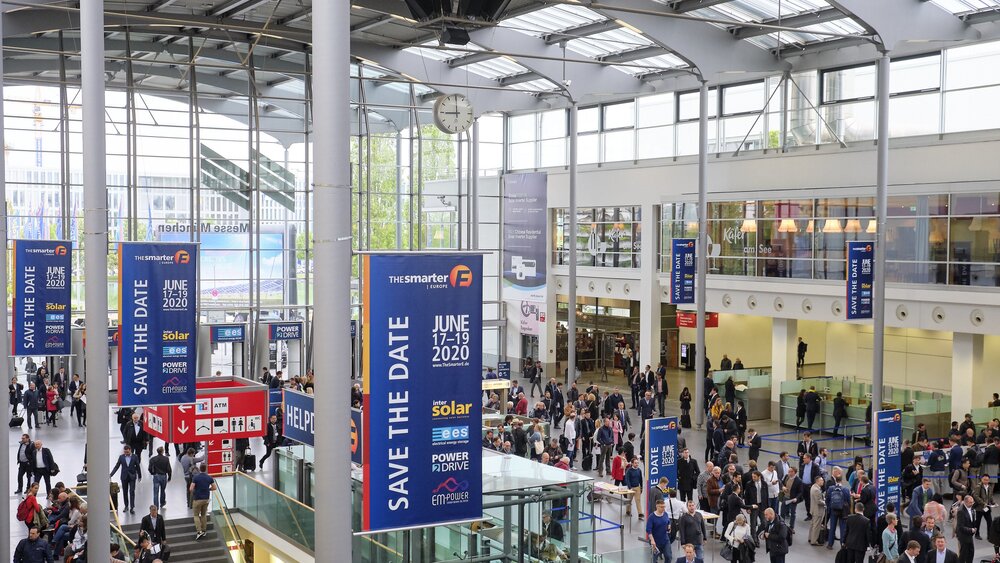A large crowd is walking through a brightly lit convention center, where banners for an event in June 2020 are on display. Glass walls, escalators, and booths are visible.