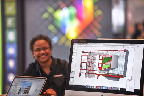 A smiling person sits behind a computer displaying colorful architecture software.