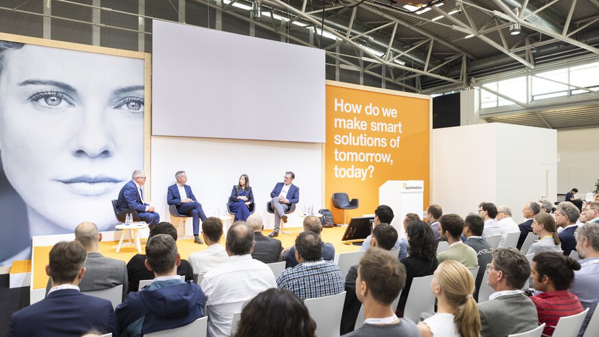 Panel discussion between 4 people on the Automatica stage. Full rows of spectators can be seen in the foreground.