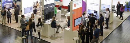 Blick von schräg oben auf mehrere Messestände der digitalBAU, an dem zahlreiche Besucher sich mit Ausstellern unterhalten.