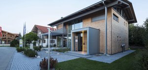 Modernes zweistöckiges Haus mit Holzpaneelen, großen Fenstern und einem schrägen Dach, umgeben von einem gepflasterten Weg und Gras.