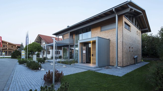 Modernes zweistöckiges Haus mit Holzpaneelen, großen Fenstern und einem schrägen Dach, umgeben von einem gepflasterten Weg und Gras.