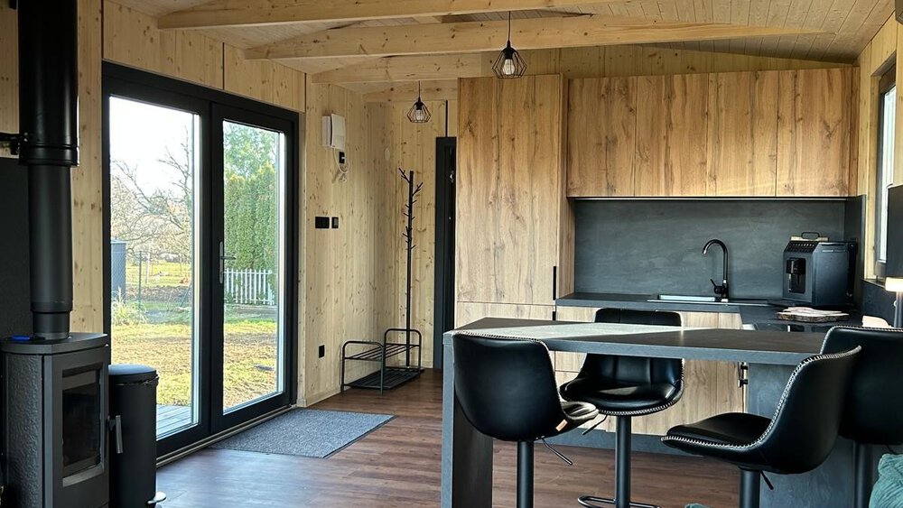 Gemütliches Hütteninterieur mit Holzakzenten, schwarzem Ofen, Barhockern an der Theke und großen Fenstern mit Blick ins Grüne.