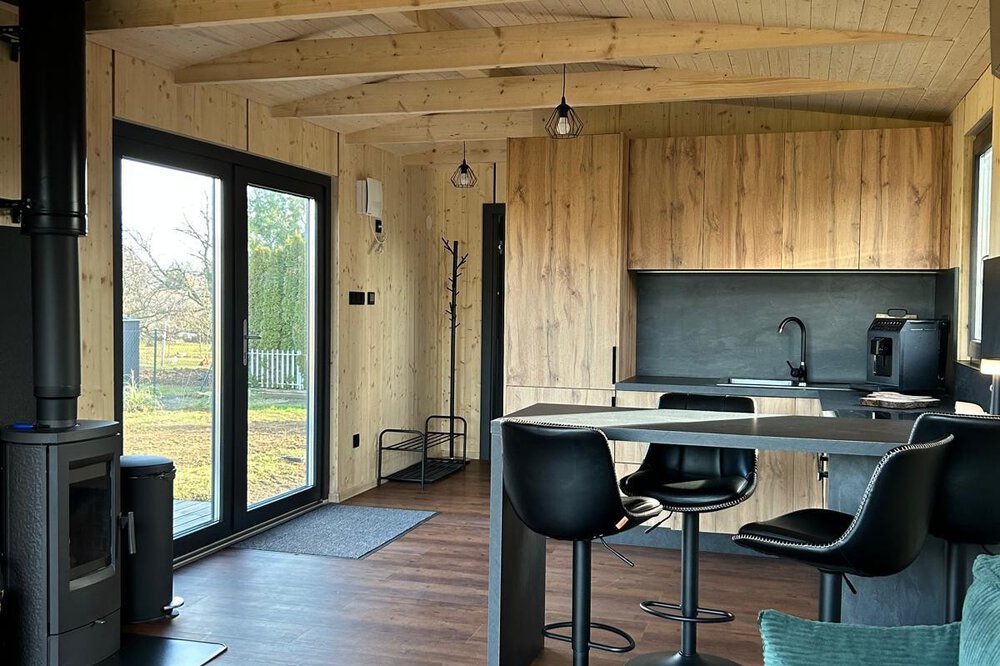 Gemütliches Hütteninterieur mit Holzakzenten, schwarzem Ofen, Barhockern an der Theke und großen Fenstern mit Blick ins Grüne.