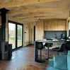 Gemütliches Hütteninterieur mit Holzakzenten, schwarzem Ofen, Barhockern an der Theke und großen Fenstern mit Blick ins Grüne.