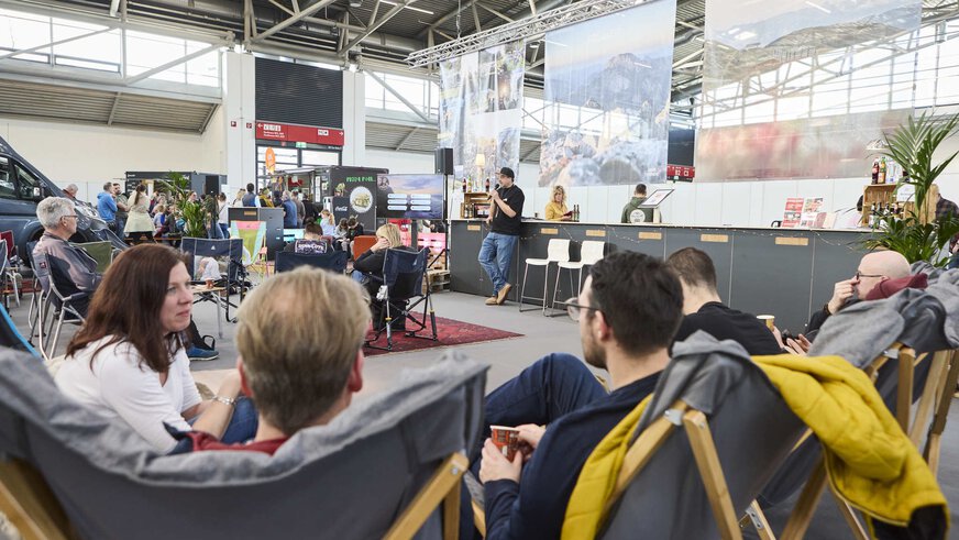 People relax in deckchairs while a speaker gives a presentation on a trade fair stage. Trade fair stands can be seen in the background.