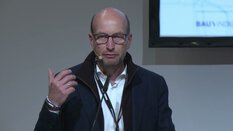 Ein Mann mit Brille und Blazer spricht an einem Rednerpult vor einem Bildschirm.