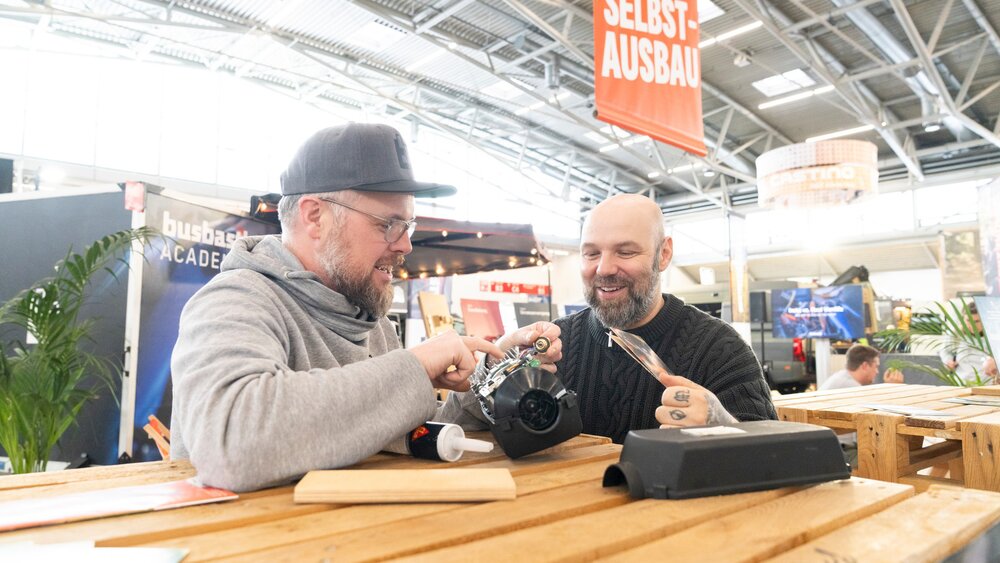 Zwei Männer, die lächelnd ein Gerät an einem Holztisch in einem hellen Veranstaltungsraum zusammenbauen.
