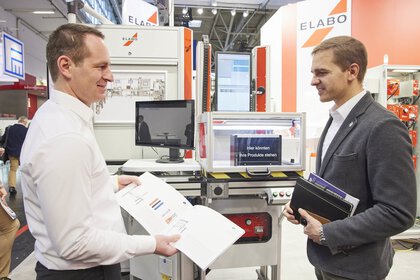 Zwei Männer in Geschäftskleidung unterhalten sich an einem Messestand auf der productronica. Sie halten Unterlagen in den Händen.