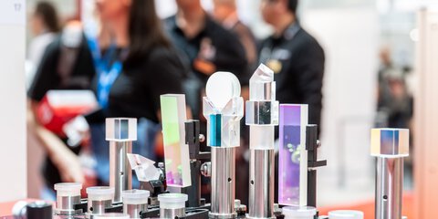 Various glass prisms and optical devices on display at a trade fair stand, people can be seen out of focus in the background.