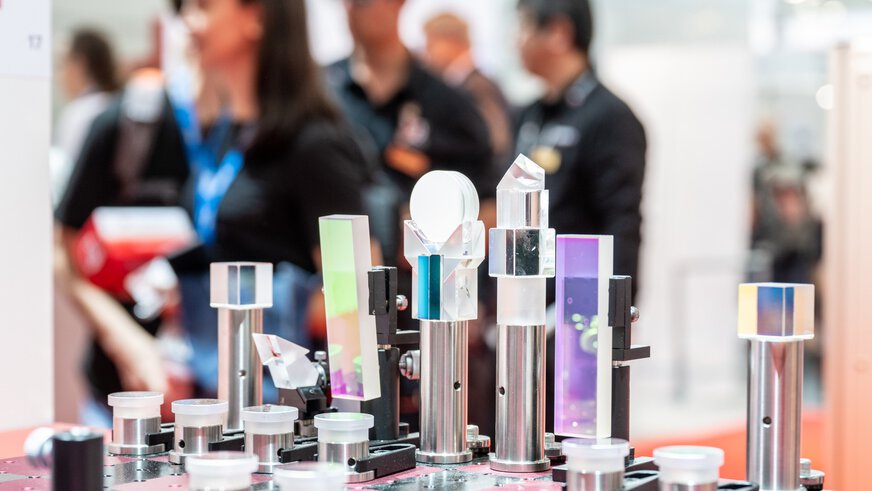 Various glass prisms and optical devices on display at a trade fair stand, people can be seen out of focus in the background.