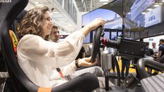 Young woman playing in a racing simulator at an exhibition stand, smiling and focused on the screen.