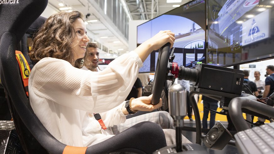 Young woman playing in a racing simulator at an exhibition stand, smiling and focused on the screen.