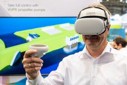 Ein Mann in einem weißen Hemd, der lächelnd ein VR-Brille verwendet. Im Hintergrund ist unscharf eine Messehalle zu sehen.
