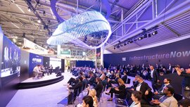 Eine Podiumsdiskussion, die auf einer Bühne im Rahmen einer großen Messe in einer Halle stattfindet, wobei das Publikum in Reihen sitzt.
