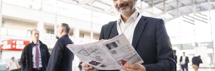 A man in a suit smiles as he reads a brochure in the bright, spacious entrance area of the LOPEC trade fair.