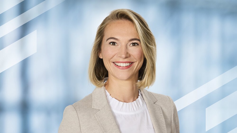 Eine lächelnde Frau mit schulterlangen blonden Haaren und Ohrsteckern trägt eine weiße Bluse und ein beiges Jackett darüber.