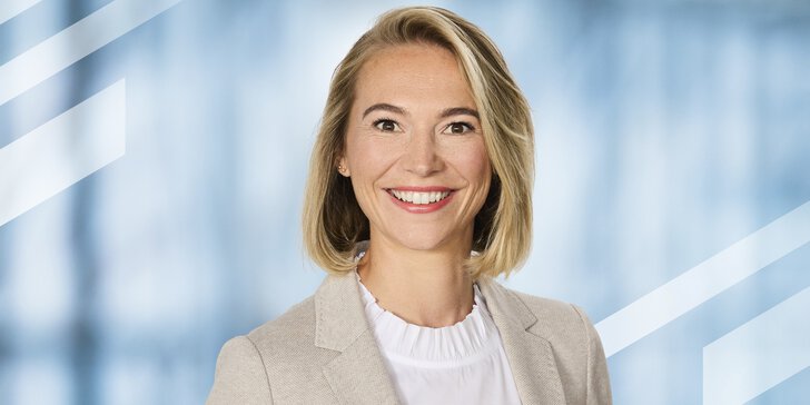 Eine lächelnde Frau mit schulterlangen blonden Haaren und Ohrsteckern trägt eine weiße Bluse und ein beiges Jackett darüber.
