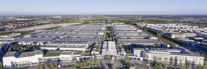 Aerial view over the Messesee, with a view of the Messe München grounds with the west entrance and the halls under a bright blue sky.