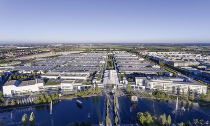 Aerial view over the Messesee, with a view of the Messe München grounds with the west entrance and the halls under a bright blue sky.