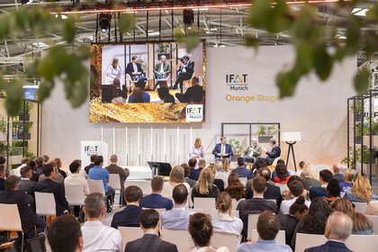 The audience is watching a panel discussion on the “Orange Stage” at IFAT Munich; the four speakers are displayed on a large screen.