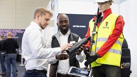 Zwei Männer stehen neben einer Schaufensterpuppe auf einem Messestand. Die Schaufensterpuppe trägt Sicherheitskleidung inklusive Helm und ein Messgerät mit der Aufschrift 