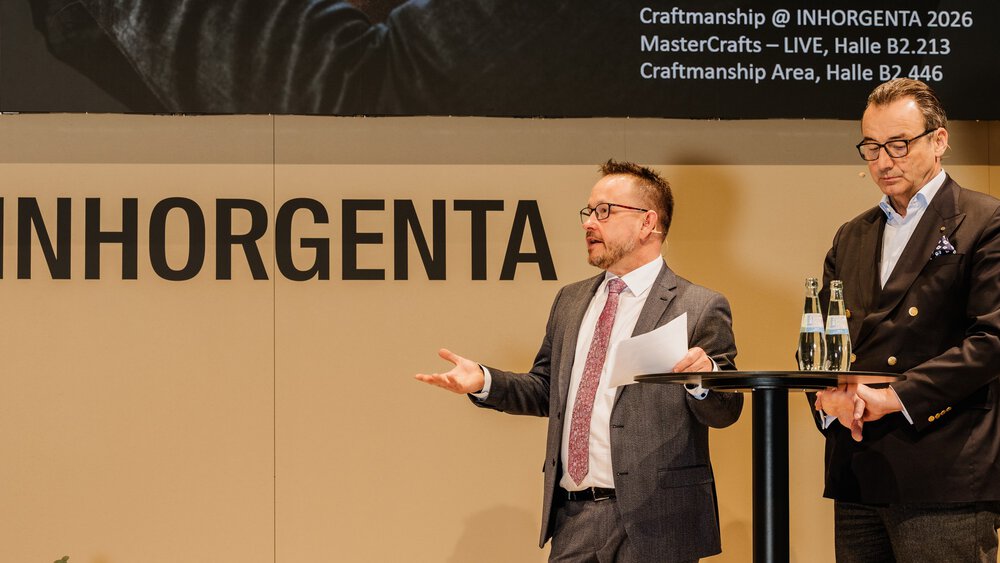Two men in suits are speaking on stage at the INHORGENTA event, standing at a high table with bottles of water.