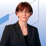 A woman with short brown hair and glasses, wearing a dark blazer, is standing in front of a blue geometric background.