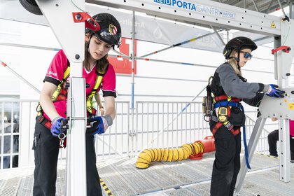 Two people in protective equipment and climbing harnesses work on scaffolding in an exhibition hall.